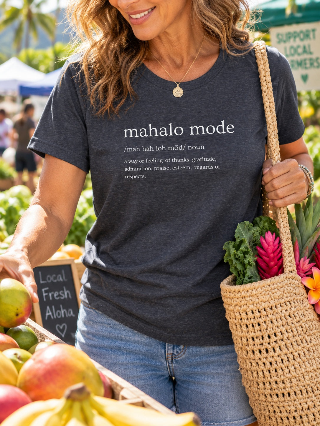 Woman wearing a charcoal Mahalo Mode definition t-shirt while shopping for fresh produce at a Hawaiʻi farmers market.