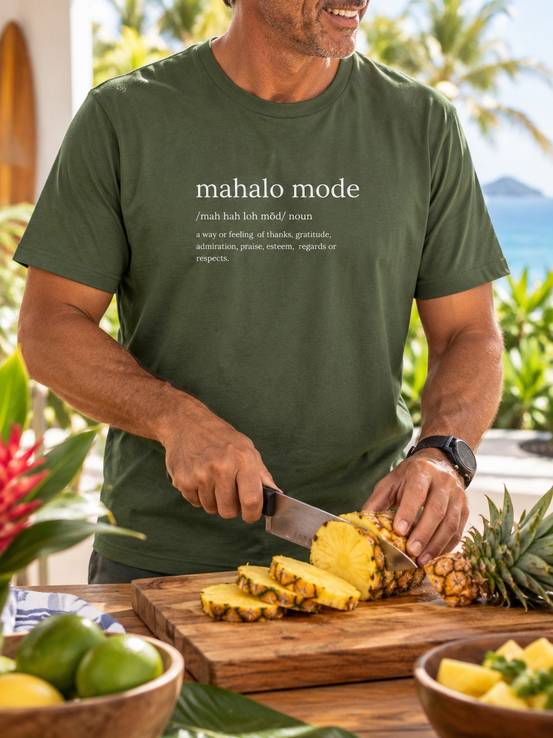 Man wearing a military green Mahalo Mode definition t-shirt while slicing fresh pineapple in a tropical Hawaiʻi kitchen.