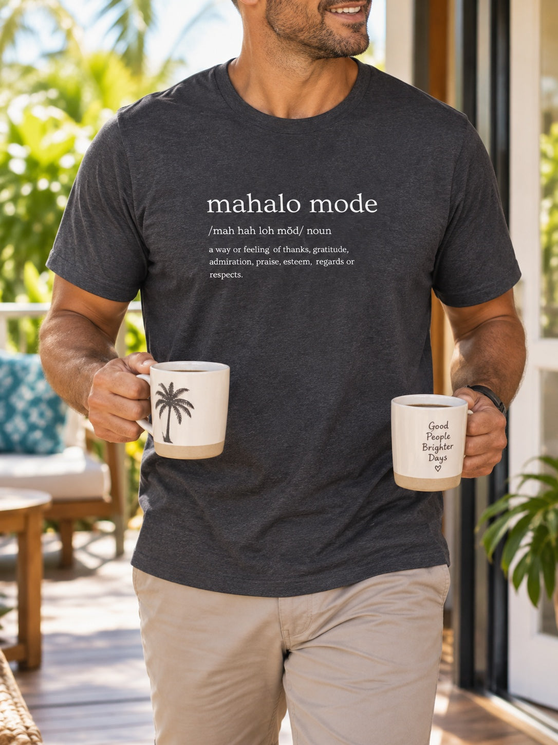 Man wearing a charcoal Mahalo Mode definition t-shirt while carrying two coffee mugs on a tropical Hawaiʻi lanai.
