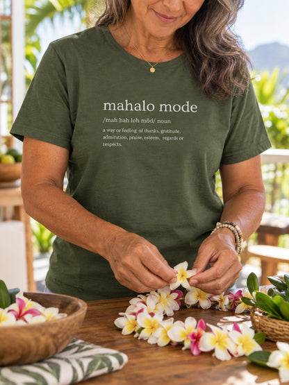 A woman wearing an olive green graphic t-shirt with the text 'mahalo mode' printed in white, shown in a tropical outdoor setting stringing plumeria flowers into a lei on a wooden table.