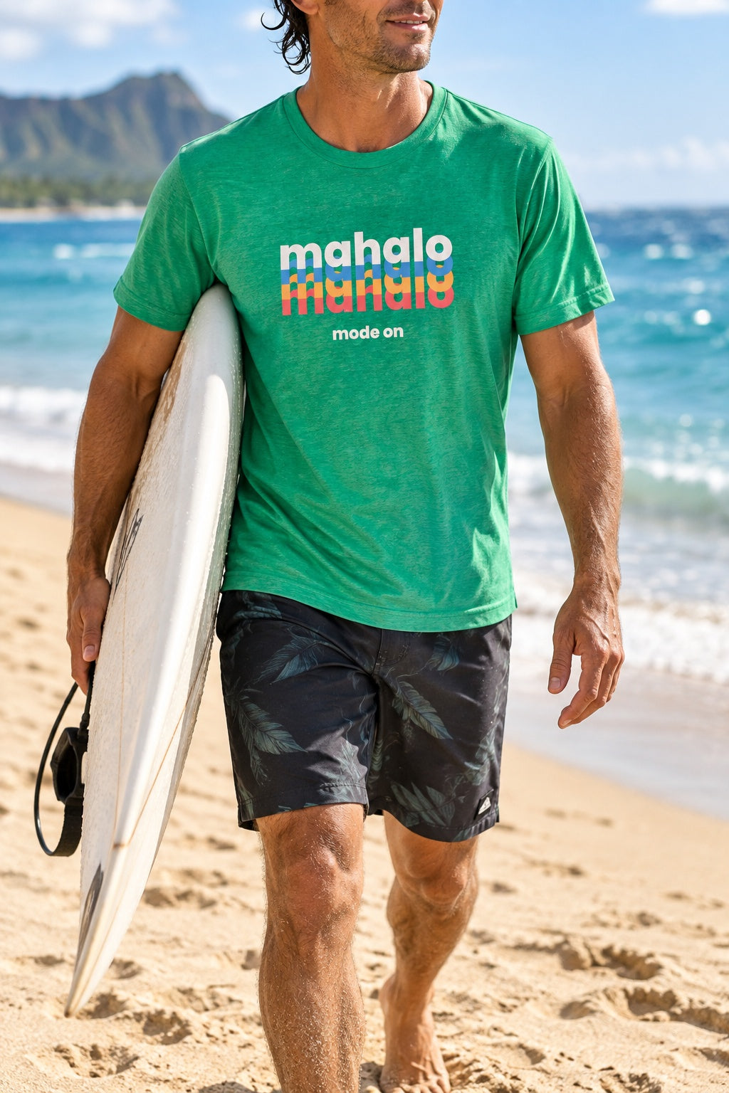 Man wearing a green retro Mahalo Mode graphic t-shirt while carrying a surfboard on a Hawaiʻi beach.