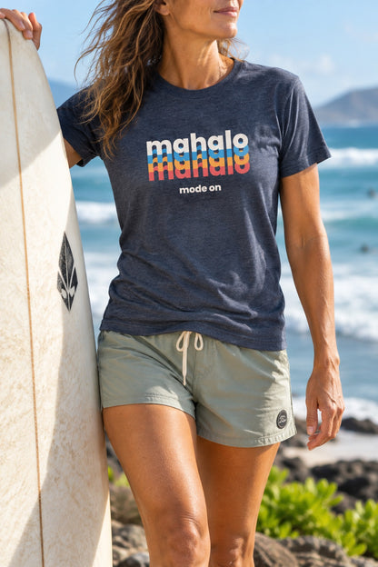 Woman wearing a heather navy retro Mahalo Mode graphic t-shirt while carrying a surfboard by the ocean in Hawaiʻi.