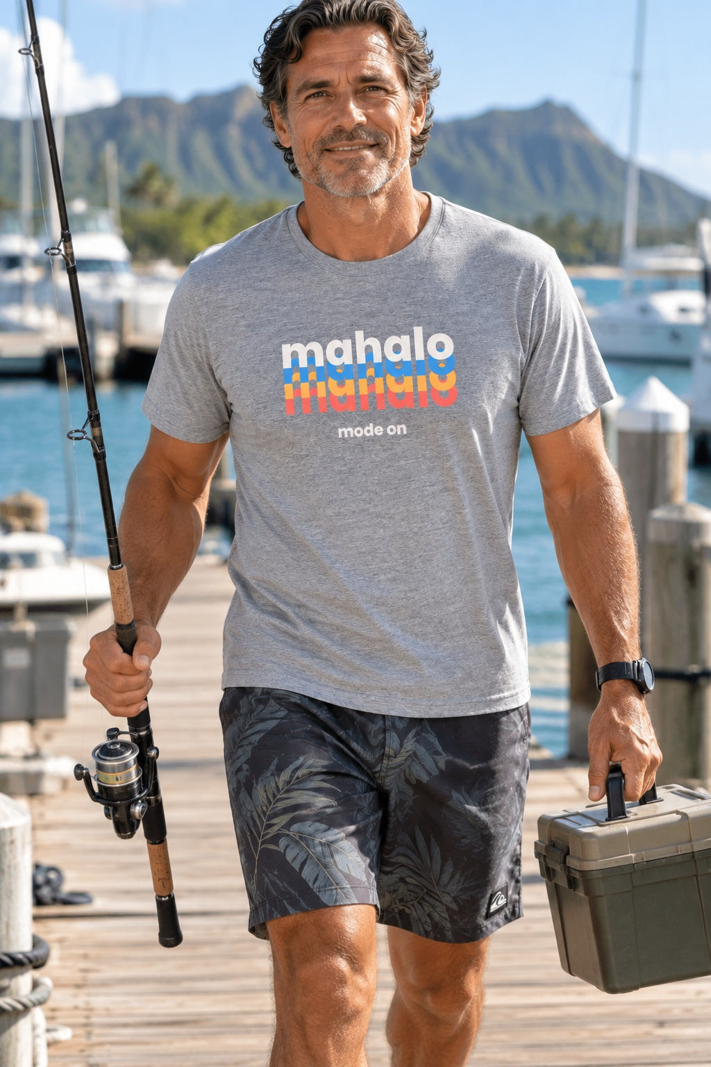 Man wearing a grey retro Mahalo Mode graphic t-shirt while carrying fishing gear at a Hawaiʻi marina.