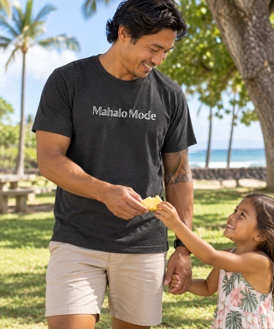 Man wearing a heather dark grey Mahalo Mode t-shirt while sharing a snack with a child at a Hawaiʻi park.