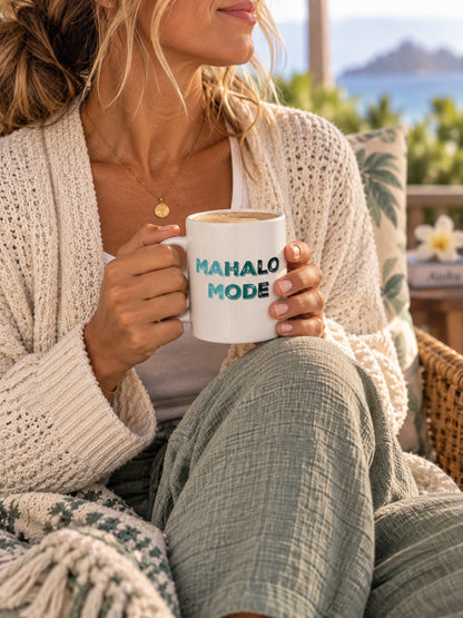A woman in a cozy knit cardigan and green pants sits outdoors holding a white ceramic mug that reads 'MAHALO MODE' in teal lettering. The background shows a sunny beach or patio setting with a pillow and distant water.
