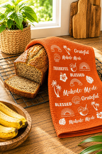 An orange kitchen towel with white tropical-themed text and illustrations, draped over a loaf of bread on a wooden table. The scene includes a sliced loaf on a cooling rack, bananas in a wooden bowl, a potted plant, and wooden cutting boards near a window.