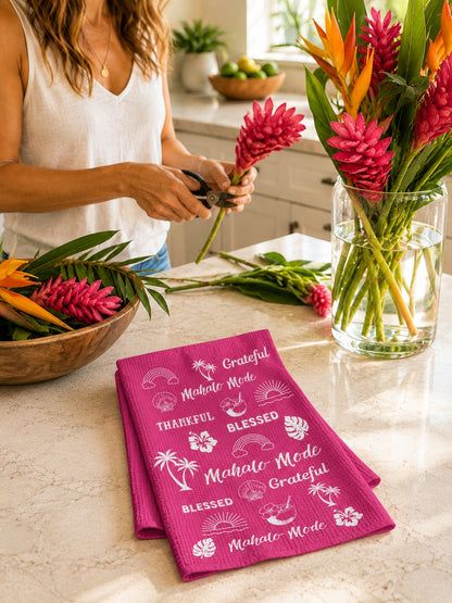 A folded bright pink kitchen towel with white tropical text and palm prints sits on a marble countertop in a kitchen. A woman in a white sleeveless top is partially visible behind the counter, holding flowers near a glass vase of colorful tropical blooms.