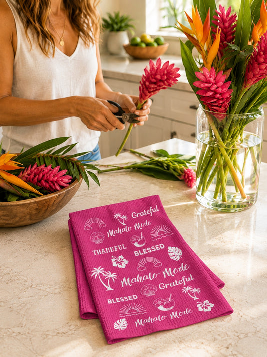 A folded bright pink kitchen towel with white tropical text and palm prints sits on a marble countertop in a kitchen. A woman in a white sleeveless top is partially visible behind the counter, holding flowers near a glass vase of colorful tropical blooms.