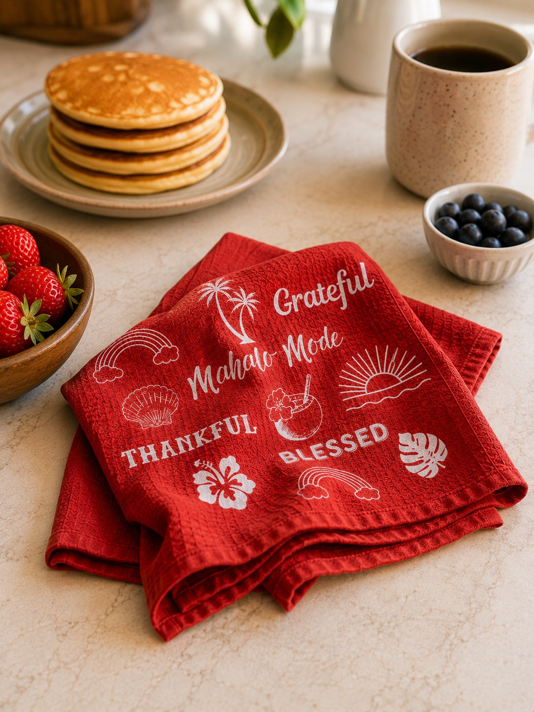 A folded red kitchen towel with white tropical and beach-themed printed text and motifs, styled on a breakfast table with pancakes, strawberries, blueberries, and a mug in the background.