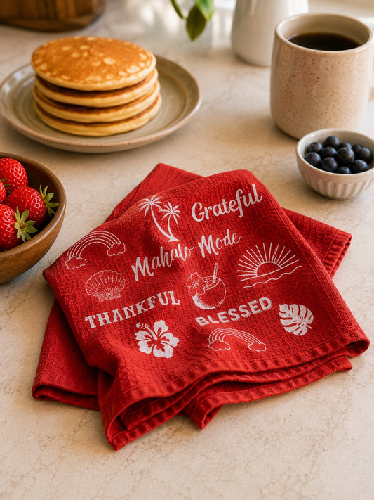 A folded red kitchen towel with white tropical and beach-themed printed text and motifs, styled on a breakfast table with pancakes, strawberries, blueberries, and a mug in the background.