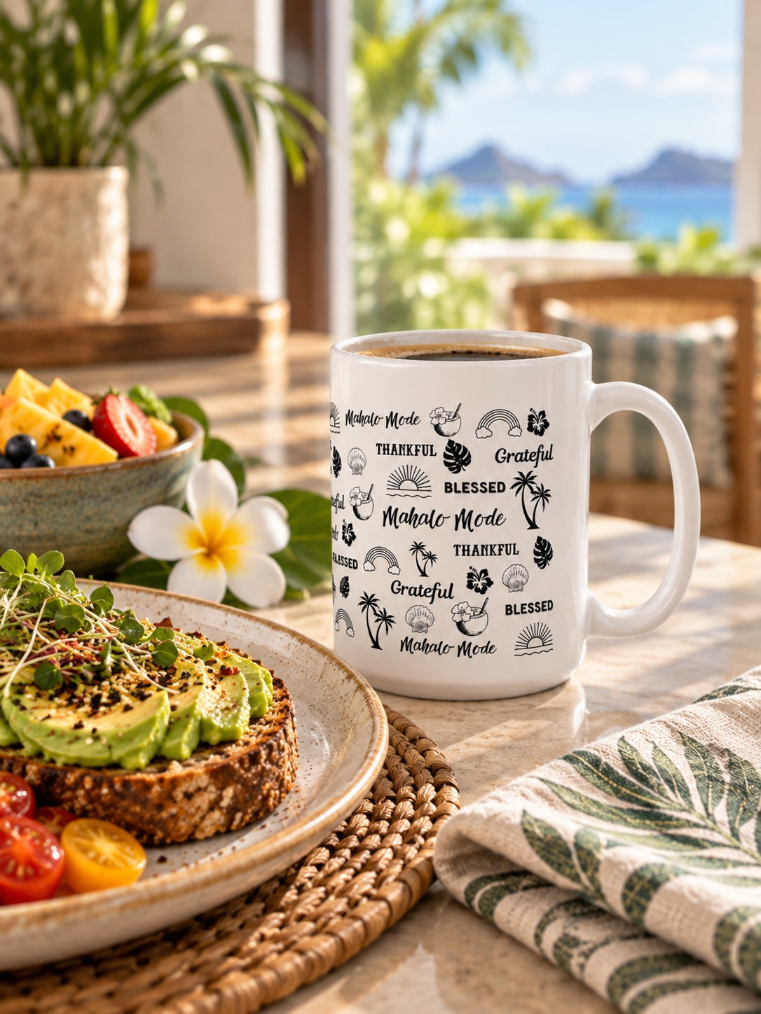 A white ceramic coffee mug with black motivational text 'mahalo mode' 'thankful' 'grateful' 'blessed' and palm tree illustrations sits on a table in a bright, tropical kitchen or cafe setting. Avocado toast, a fruit salad, and a striped cloth napkin are nearby, with a scenic ocean view in the background.