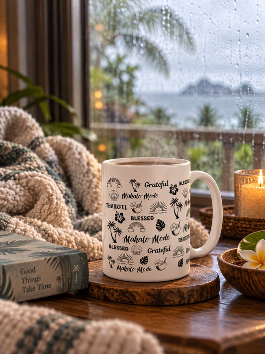 A white ceramic mug with black tropical-themed text and illustrations 'mahalo mode' 'thankful' 'grateful' 'blessed' sits on a wooden coaster on a table by a window. A cozy knit blanket, a book, a lit candle, and a flower are arranged around it, with a rainy outdoor scene visible in the background.
