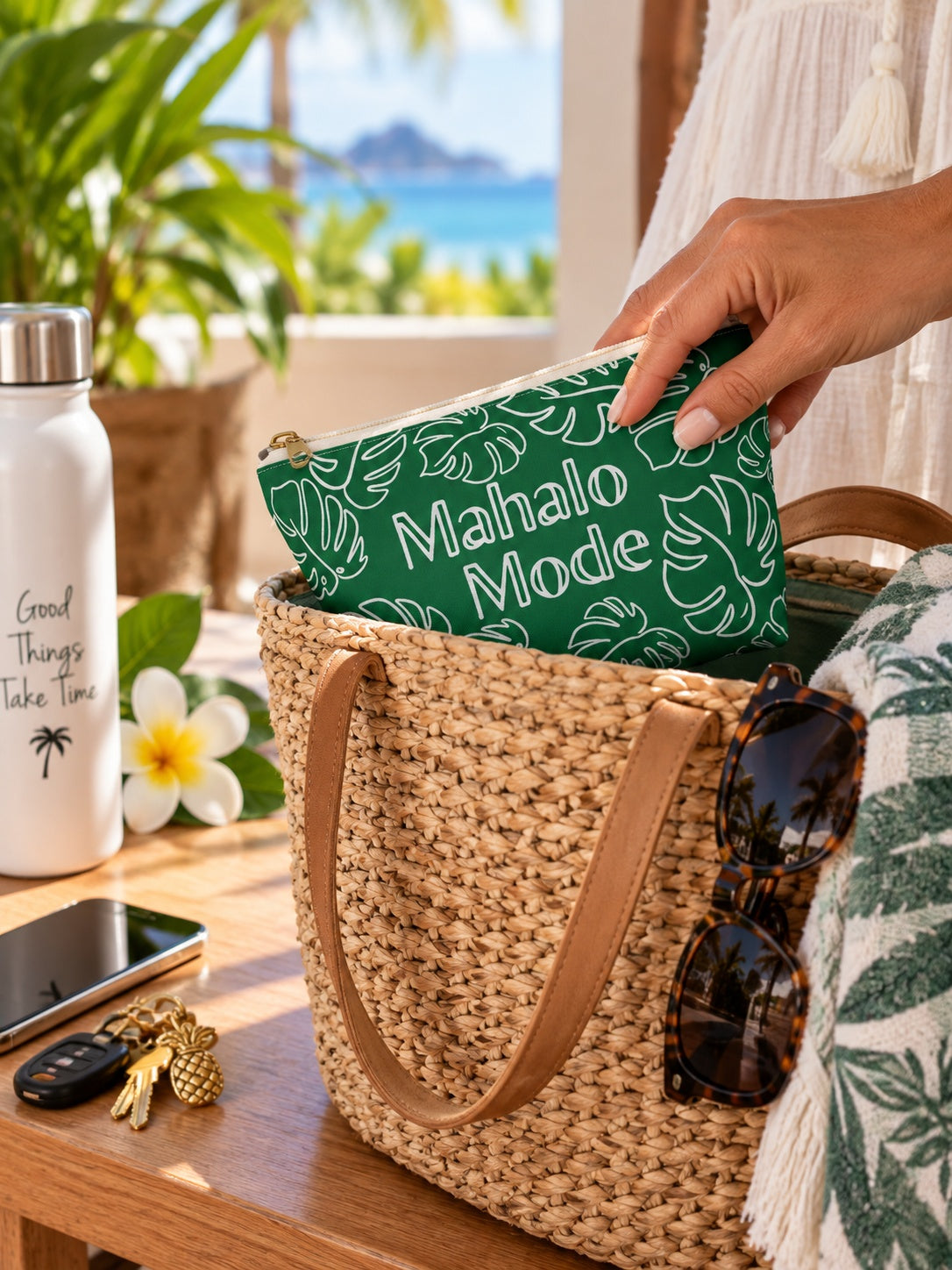 A woven straw tote bag sits on a wooden table with a hand holding a green zip pouch printed with the words 'Mahalo Mode' in white. The scene includes sunglasses, a white bottle, keys, a phone, and tropical beach scenery in the background.