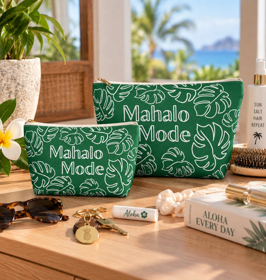 Two green zip pouches with white monstera leaf prints and the words 'Mahalo Mode' are displayed on a wooden table in a tropical beach setting. The scene includes sunglasses, a lip balm, a book, and a plant in the background.