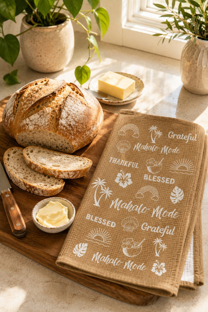 A rustic kitchen scene with a loaf of bread, sliced bread, butter, and a beige tan tea towel printed with white tropical motifs and phrases like 'Grateful,' 'Blessed,' and 'Mahalo Mode.' The towel is styled on a wooden board beside the bread, with plants and a window in the background.