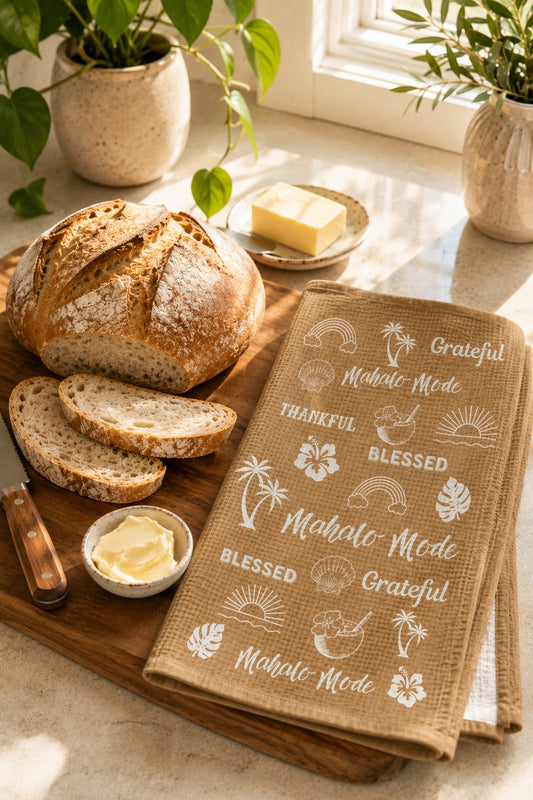 A rustic kitchen scene with a loaf of bread, sliced bread, butter, and a beige tan tea towel printed with white tropical motifs and phrases like 'Grateful,' 'Blessed,' and 'Mahalo Mode.' The towel is styled on a wooden board beside the bread, with plants and a window in the background.