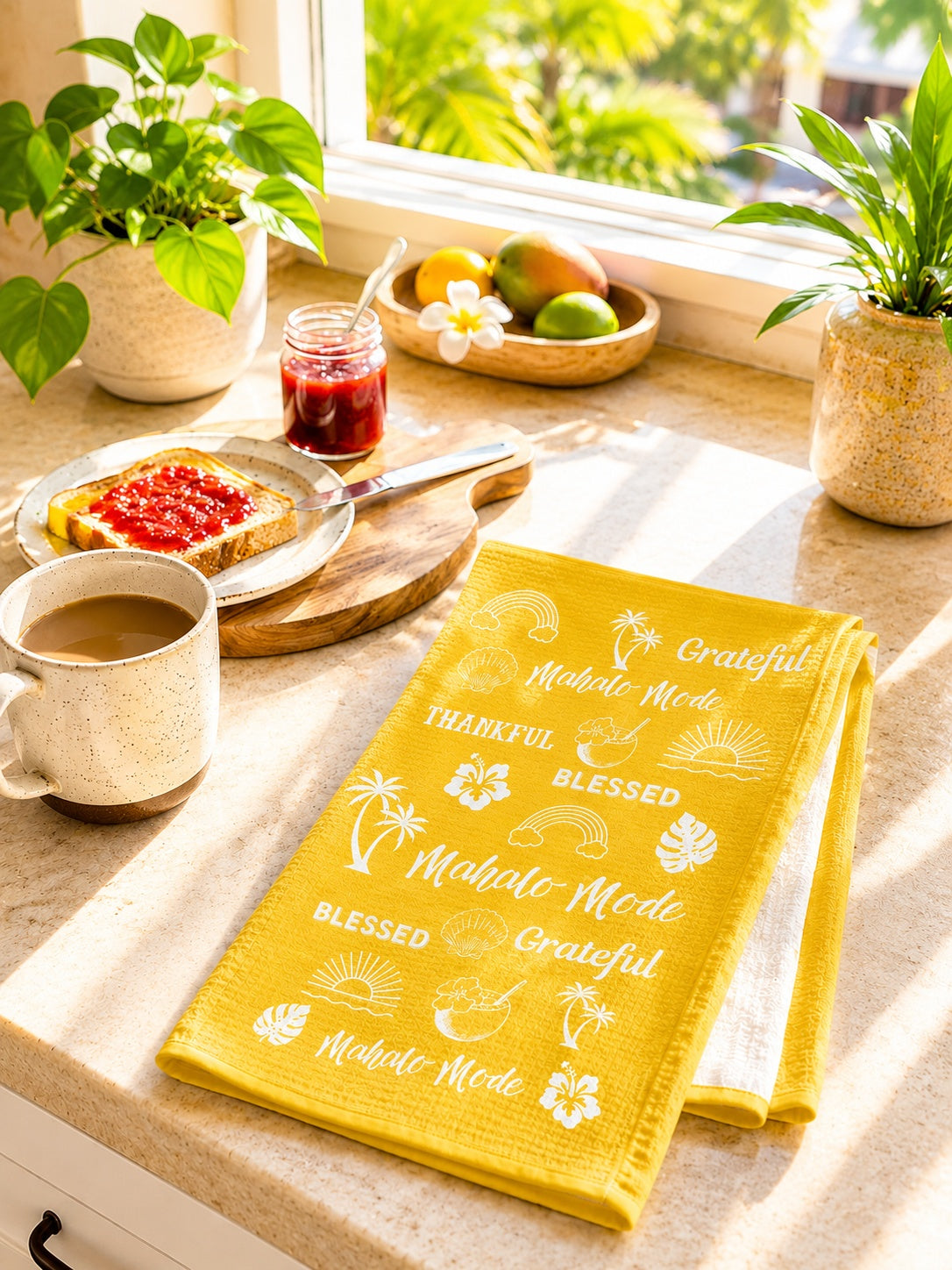 A folded yellow kitchen towel with white Hawaiian-themed text and illustrations, including 'Mahalo Mode' and 'Grateful', displayed on a kitchen counter next to a mug of coffee, toast with jam, and plants by a window.