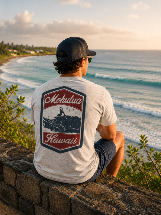 A man sits on a stone wall overlooking the ocean, viewed from behind. He is wearing a light gray t-shirt with a large red, navy, and white graphic on the back that reads 'Mokulua Hawaii' with an illustration of a surfer, along with a black baseball cap and dark shorts.