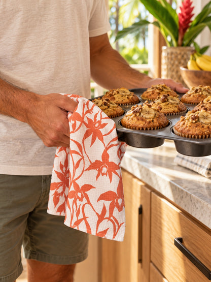 Man using an Orchid Pumpkin kitchen towel to hold a tray of freshly baked banana muffins in a tropical Hawaiʻi kitchen.