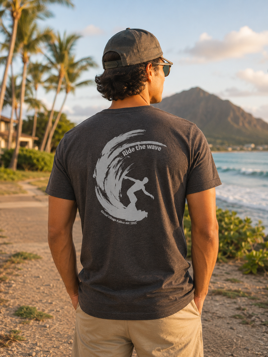 A man standing on a beach path, viewed from behind, wearing a dark charcoal gray t-shirt with a large white graphic of a surfer riding a wave on the back. The background features palm trees, ocean waves, and a mountain under a warm sunset sky.