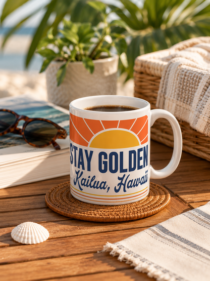 A white ceramic mug with a retro sunset design and the text 'STAY GOLDEN Kailua, Hawaii' sits on a woven coaster on a wooden table. The scene is styled with a beach-themed background including sunglasses, a book, a wicker basket, a towel, and a seashell.
