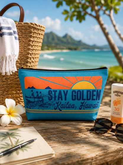 A blue zippered pouch with a retro sunset graphic reading 'STAY GOLDEN Kailua, Hawaii' sits on a wooden table outdoors by the beach. A woven tote, towel, notebook, sunglasses, and sunscreen are arranged around it, with a tropical ocean and mountain background.