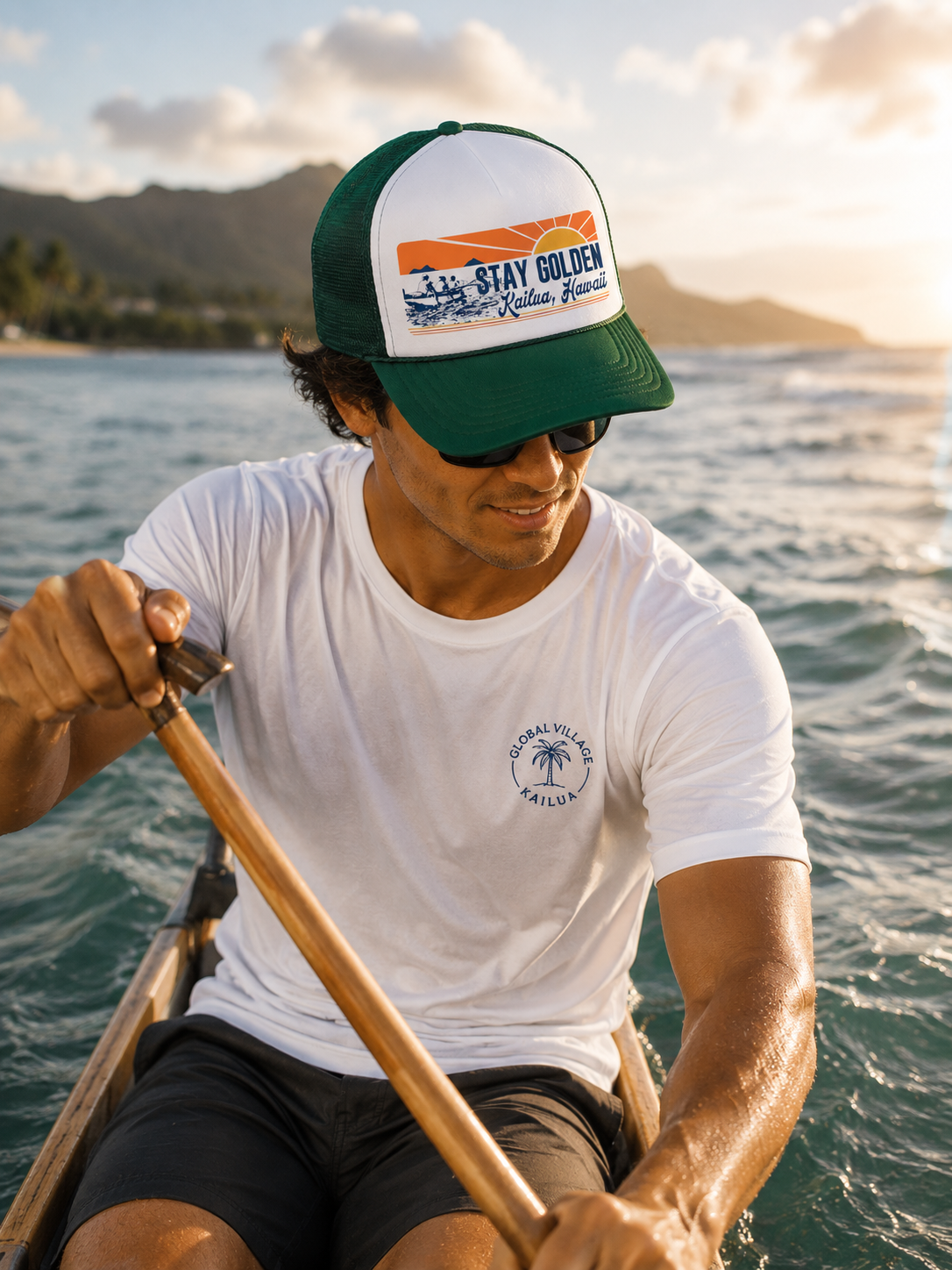 A man in a boat wearing a green and white trucker hat with a 'Stay Golden graphic and a white t-shirt with a circular logo, paddling on the water during sunset.