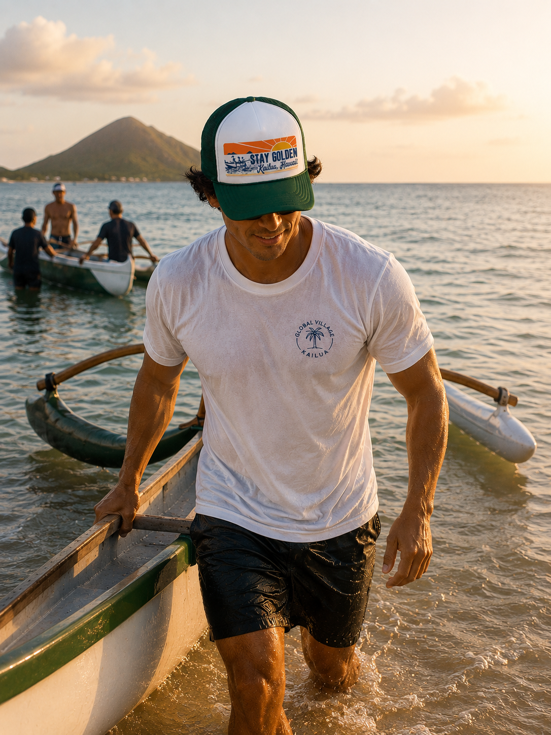 A man bringing his canoe out of the ocean, wearing a white t-shirt with a small circular logo on the chest, black shorts, and a green and white trucker hat that reads 'Stay Golden'. In the background, other boats and a mountain are visible under a warm sunset sky.