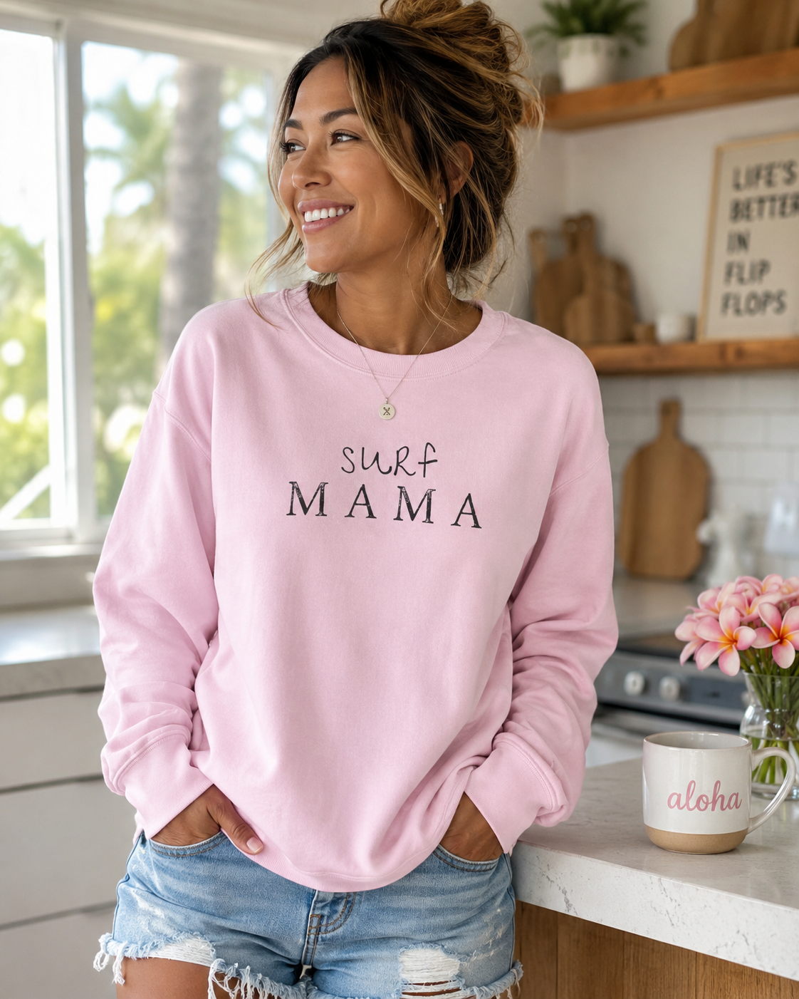 Woman wearing a light pink Surf Mama crewneck sweatshirt in a bright kitchen, featuring an original Hawaii-inspired graphic by Global Village Kailua.