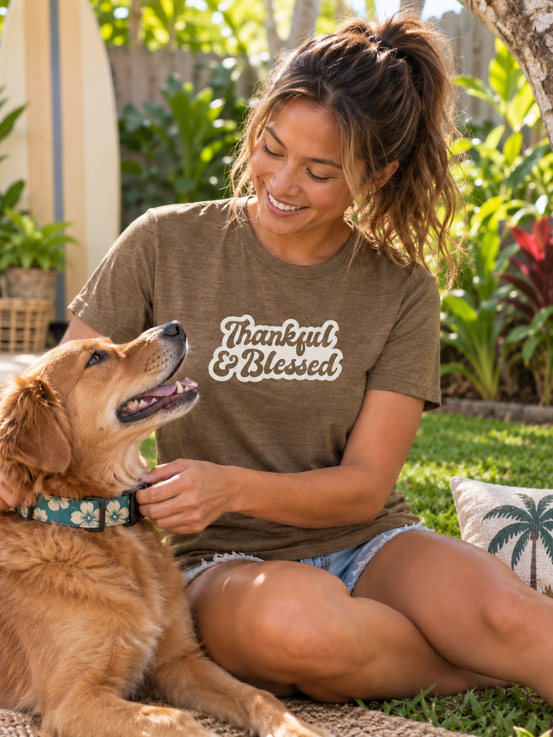 A woman sitting outdoors on the grass with a golden retriever dog, wearing a brown t-shirt with the text 'Thankful & Blessed' in a cream script. The shirt has a distressed, washed texture.