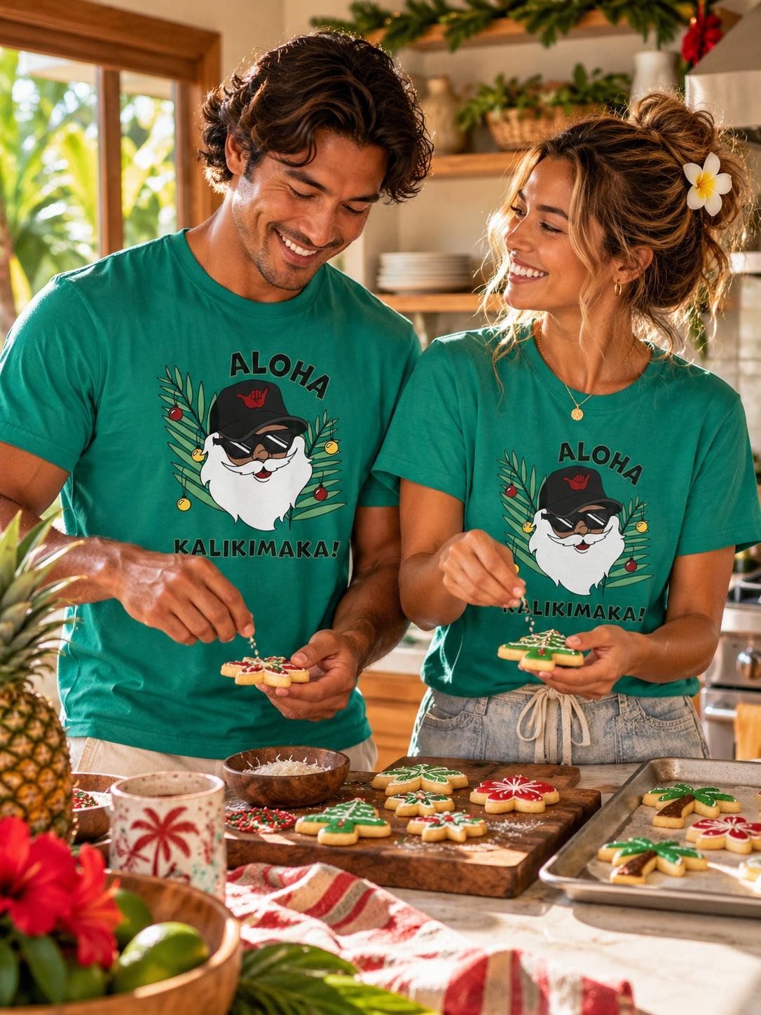 Couple wearing green Aloha Kalikimaka Santa graphic tees decorating Christmas cookies in a tropical kitchen.