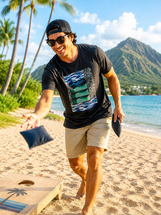 A man wearing a black graphic t-shirt with waves in motion graphic, beige shorts, and a black cap plays cornhole on a sandy beach with a tropical mountain and ocean background.