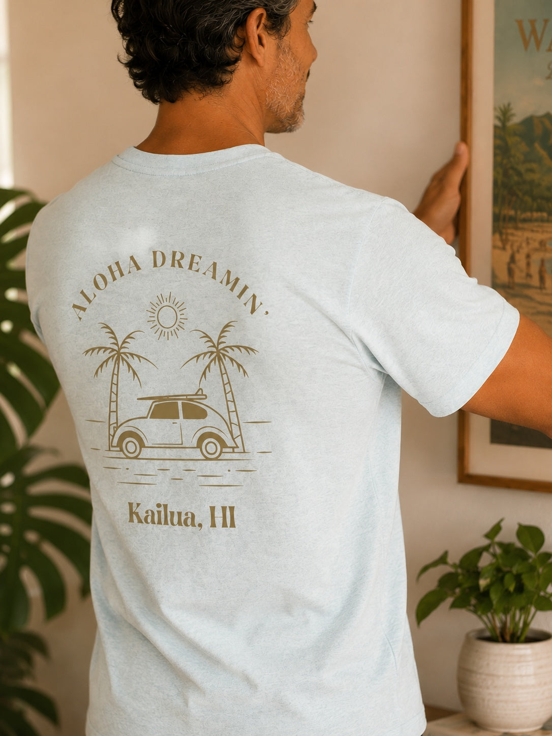 A man wearing a light heather blue t-shirt with a brown graphic print on the back, featuring the text 'ALOHA DREAMIN'' and 'Kailua, HI' with a car and palm trees, in a warm indoor setting.