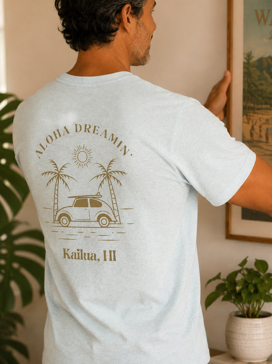 A man wearing a light heather blue t-shirt with a brown graphic print on the back, featuring the text 'ALOHA DREAMIN'' and 'Kailua, HI' with a car and palm trees, in a warm indoor setting.