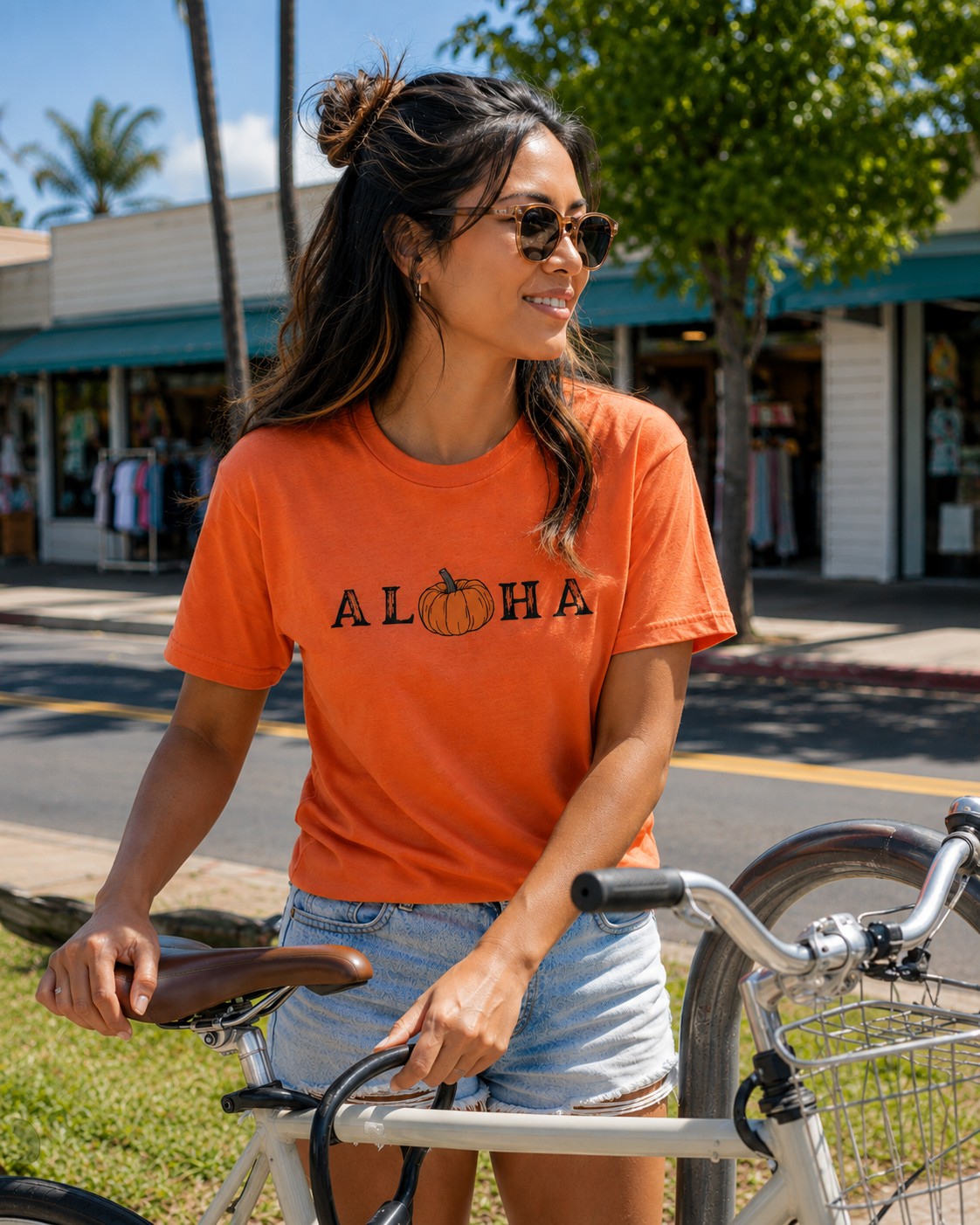 A woman wearing a bright orange graphic t-shirt with 'ALOHA' and a pumpkin design, paired with denim shorts and sunglasses, posing with a bicycle outdoors on a sunny street.
