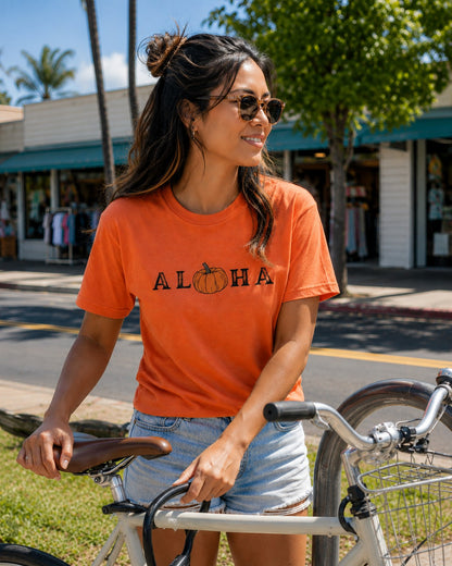 A woman wearing a bright orange graphic t-shirt with 'ALOHA' and a pumpkin design, paired with denim shorts and sunglasses, posing with a bicycle outdoors on a sunny street.
