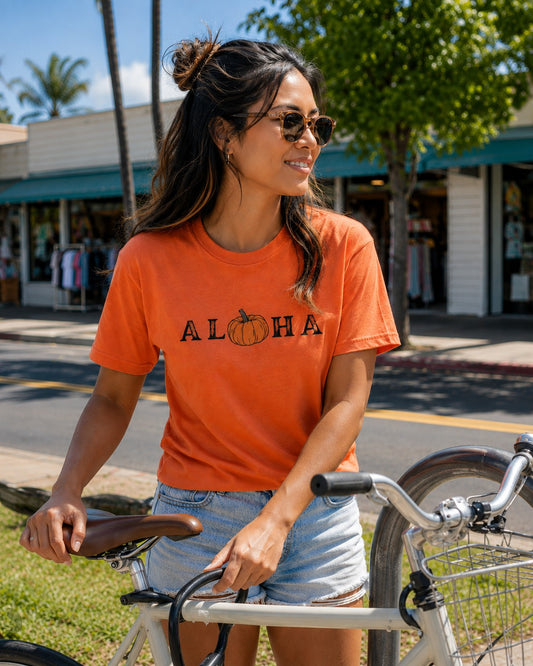 A woman wearing a bright orange graphic t-shirt with 'ALOHA' and a pumpkin design, paired with denim shorts and sunglasses, posing with a bicycle outdoors on a sunny street.