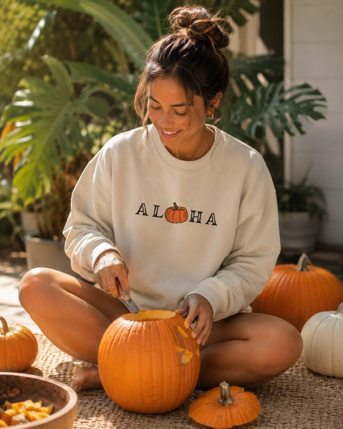 A woman sits cross-legged on a woven rug carving a pumpkin outdoors, wearing a cream sweatshirt with 'ALOHA' and a small pumpkin graphic printed across the chest. Orange pumpkins and green plants are visible in the background.