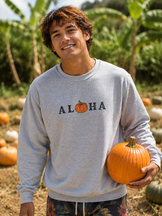 A smiling man in a pumpkin patch wearing a light gray crewneck sweatshirt with 'ALOHA' and a small pumpkin graphic on the chest, holding a large orange pumpkin.