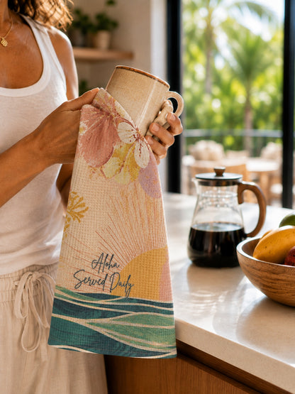 A woman in a white tank top holds a ceramic mug wrapped in a decorative kitchen towel with a floral and sunburst design. The towel reads 'Alia Served Daily' and features pink, yellow, teal, and beige tones.