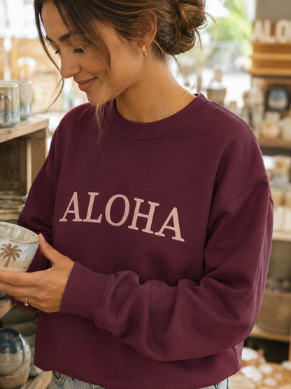 A woman wearing a maroon crewneck sweatshirt with 'ALOHA' printed in light pink across the chest, holding a ceramic mug in a cozy, softly blurred shop setting.