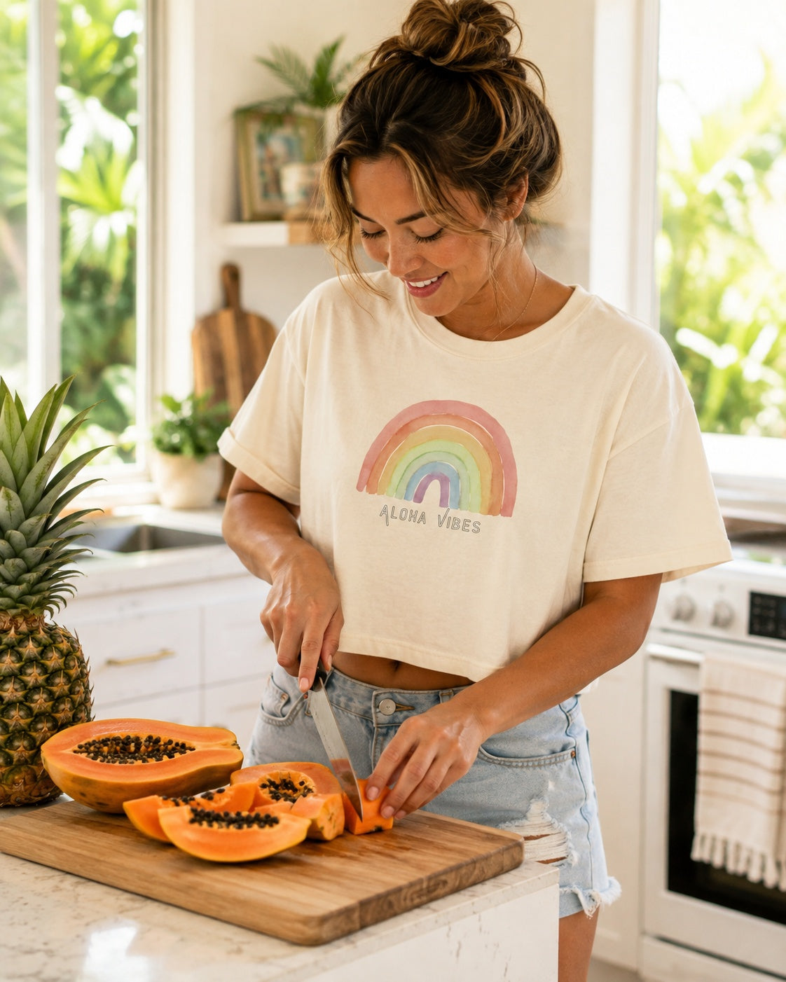 A woman in a kitchen wearing a cream-colored cropped t-shirt with a colorful rainbow graphic and the text 'ALOHA VIBES'. She is cutting papayas on a wooden cutting board next to a pineapple.