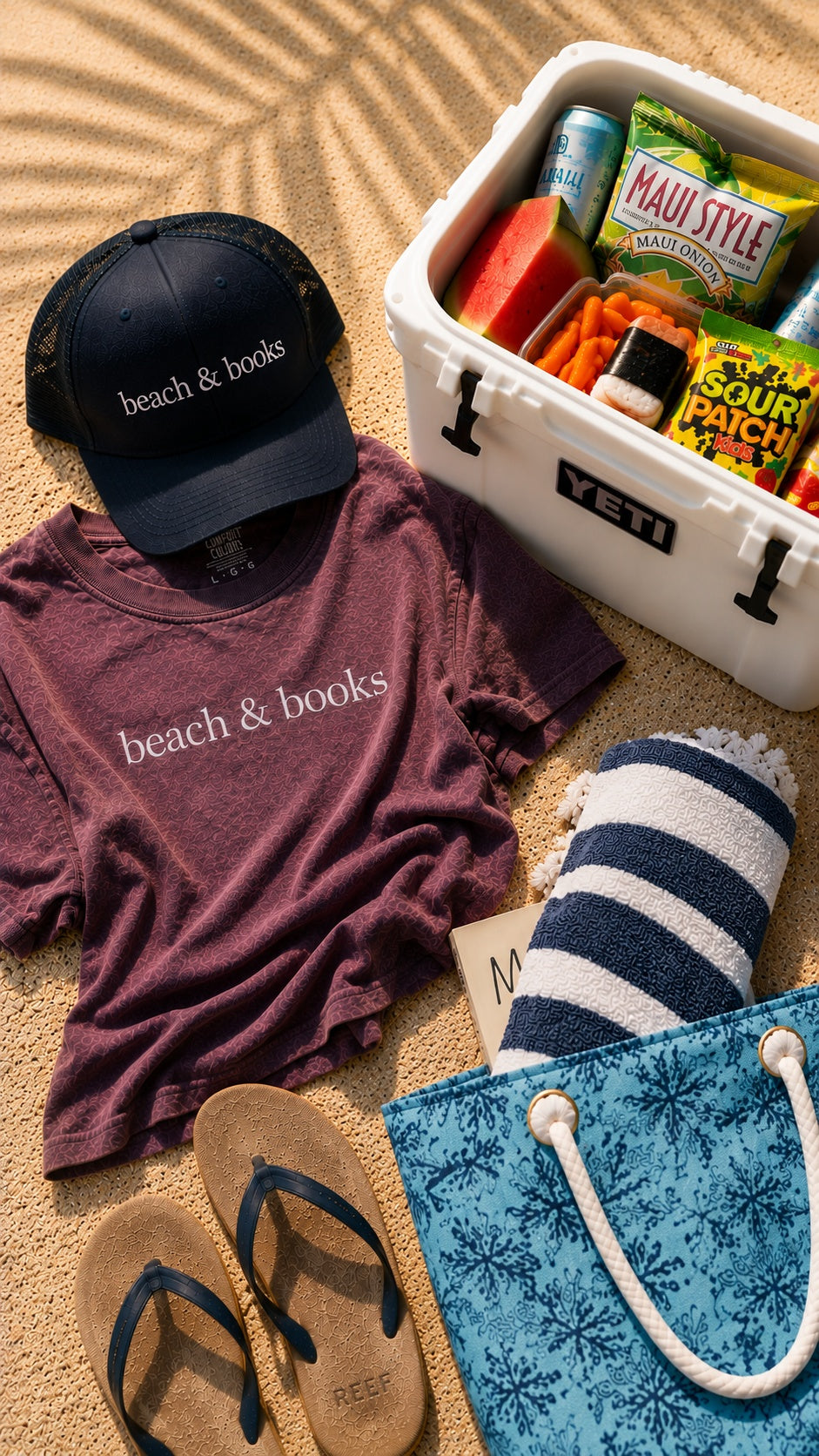 A styled beach-themed flat lay on sand featuring a dark navy cap and heathered mauve t-shirt both printed with 'beach & books', a white YETI cooler filled with snacks, a blue-and-white striped towel, a blue patterned tote bag, and tan flip-flops.