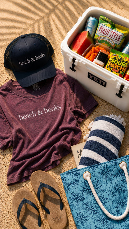 A styled beach-themed flat lay on sand featuring a dark navy cap and heathered mauve t-shirt both printed with 'beach & books', a white YETI cooler filled with snacks, a blue-and-white striped towel, a blue patterned tote bag, and tan flip-flops.