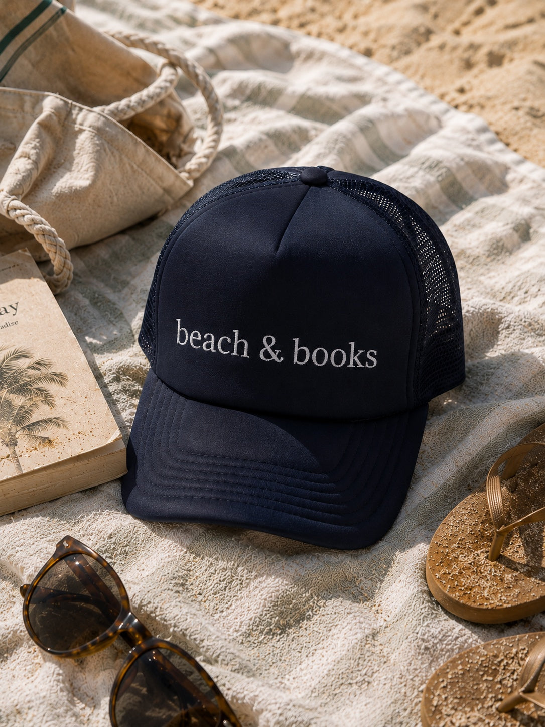 A navy blue trucker hat with 'beach & books' embroidered in white on the front, styled on a striped beach blanket with sunglasses, a book, a tote bag, and flip-flops on sand.