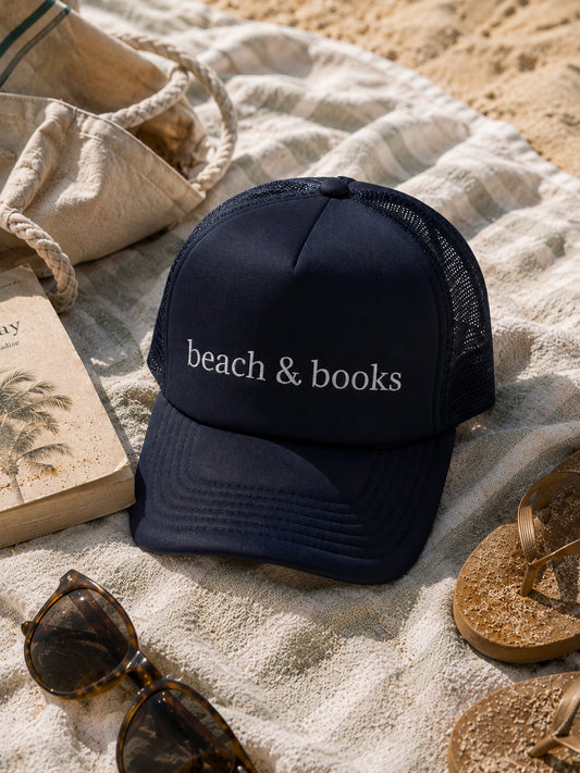 A navy blue trucker hat with 'beach & books' embroidered in white on the front, styled on a striped beach blanket with sunglasses, a book, a tote bag, and flip-flops on sand.