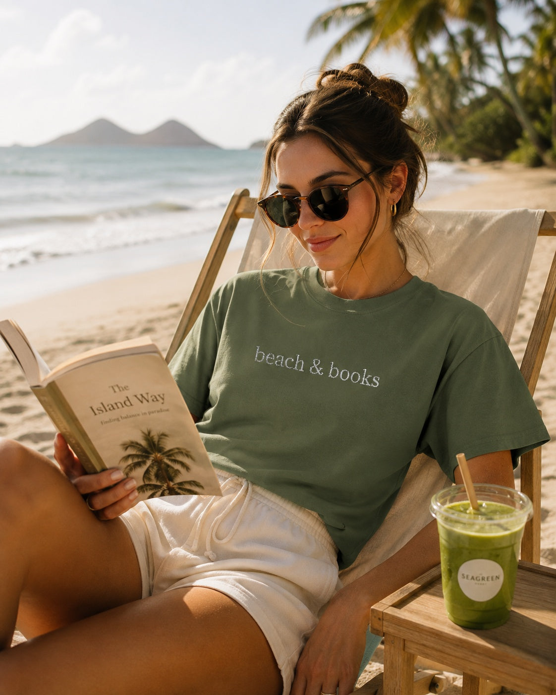 A woman sitting in a beach chair on a sandy beach, wearing a sage green t-shirt with the words 'beach & books' printed in white. She is holding an open book and wearing white shorts, with a green drink on a wooden table next to her.