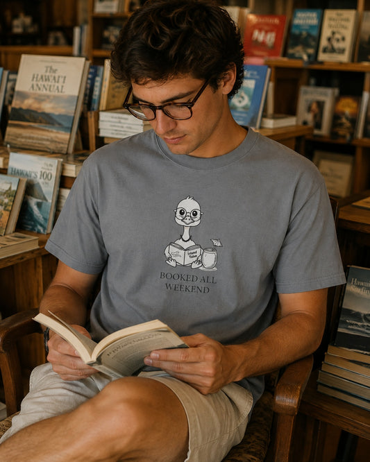 A man wearing a gray graphic t-shirt and beige shorts sits in a wooden chair inside a bookshop, reading an open book. The t-shirt features a cartoon nene goose holding a book and the text 'BOOKED ALL WEEKEND'.