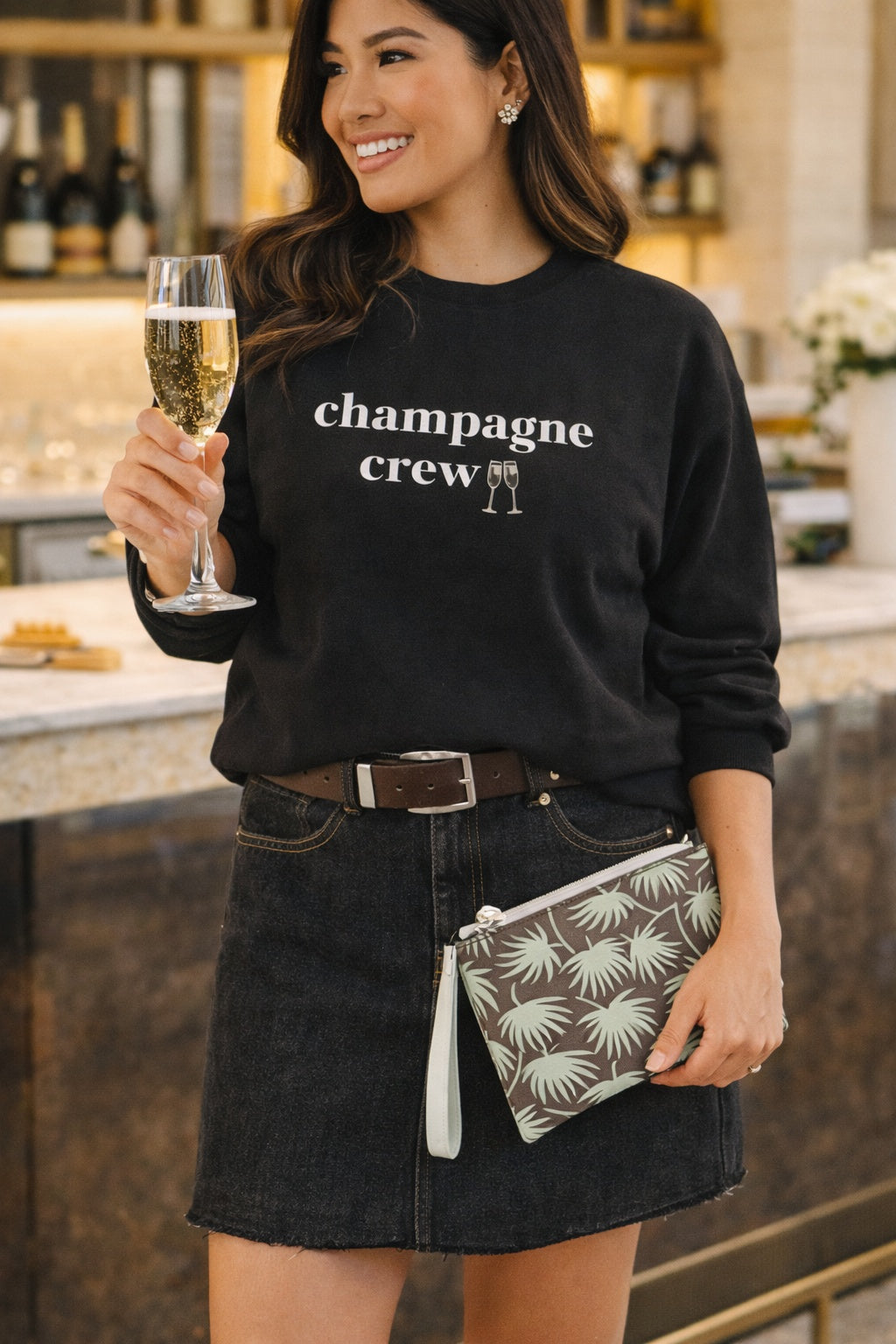 A woman wearing a black crewneck sweatshirt with 'champagne crew' printed in white text and a small champagne glass icon, paired with a dark denim mini skirt and holding a patterned clutch and a glass of champagne in a kitchen setting.