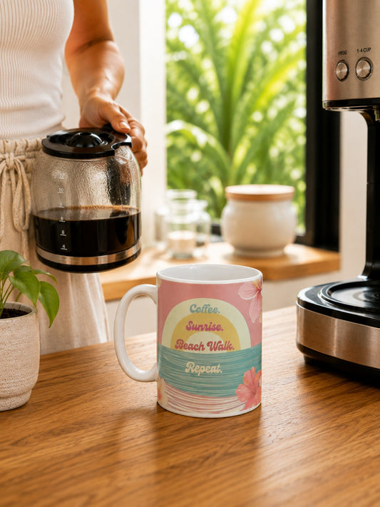 A ceramic coffee mug with a retro sunset beach graphic sits on a wooden table in a kitchen setting, with a person pouring coffee from a glass carafe in the background.